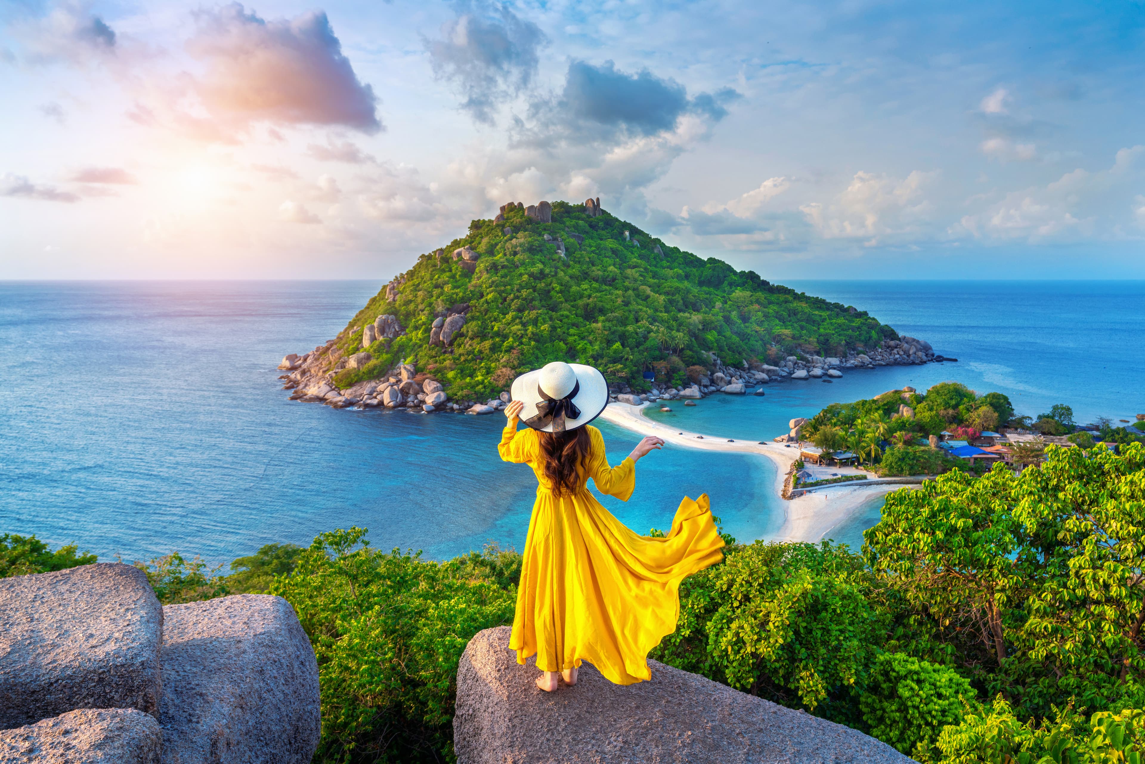 Woman in yellow dress overlooks a tropical island and sandbar from a scenic rocky viewpoint.