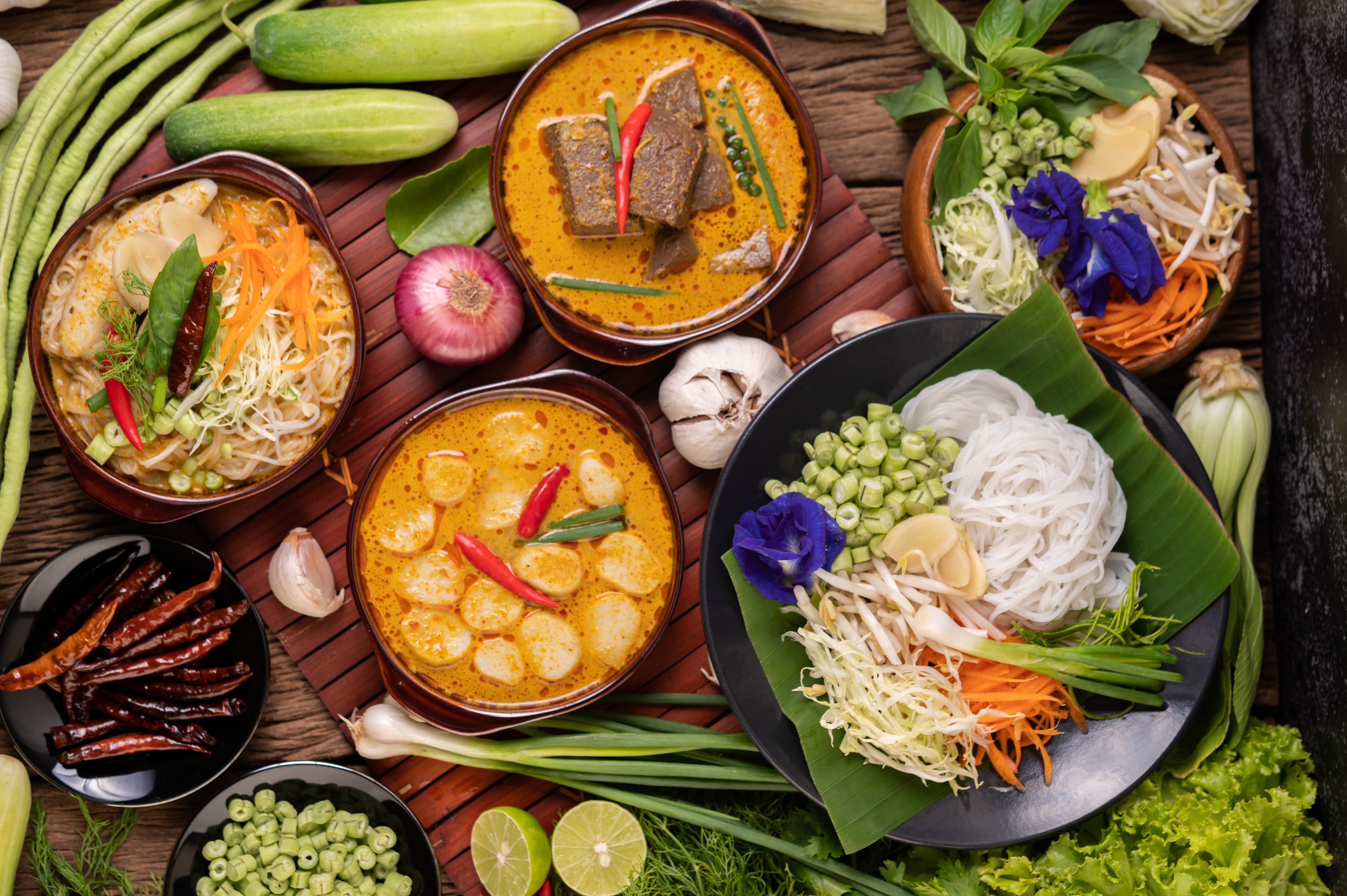 Thai rice noodles with various curries and fresh vegetables on a rustic wooden table.