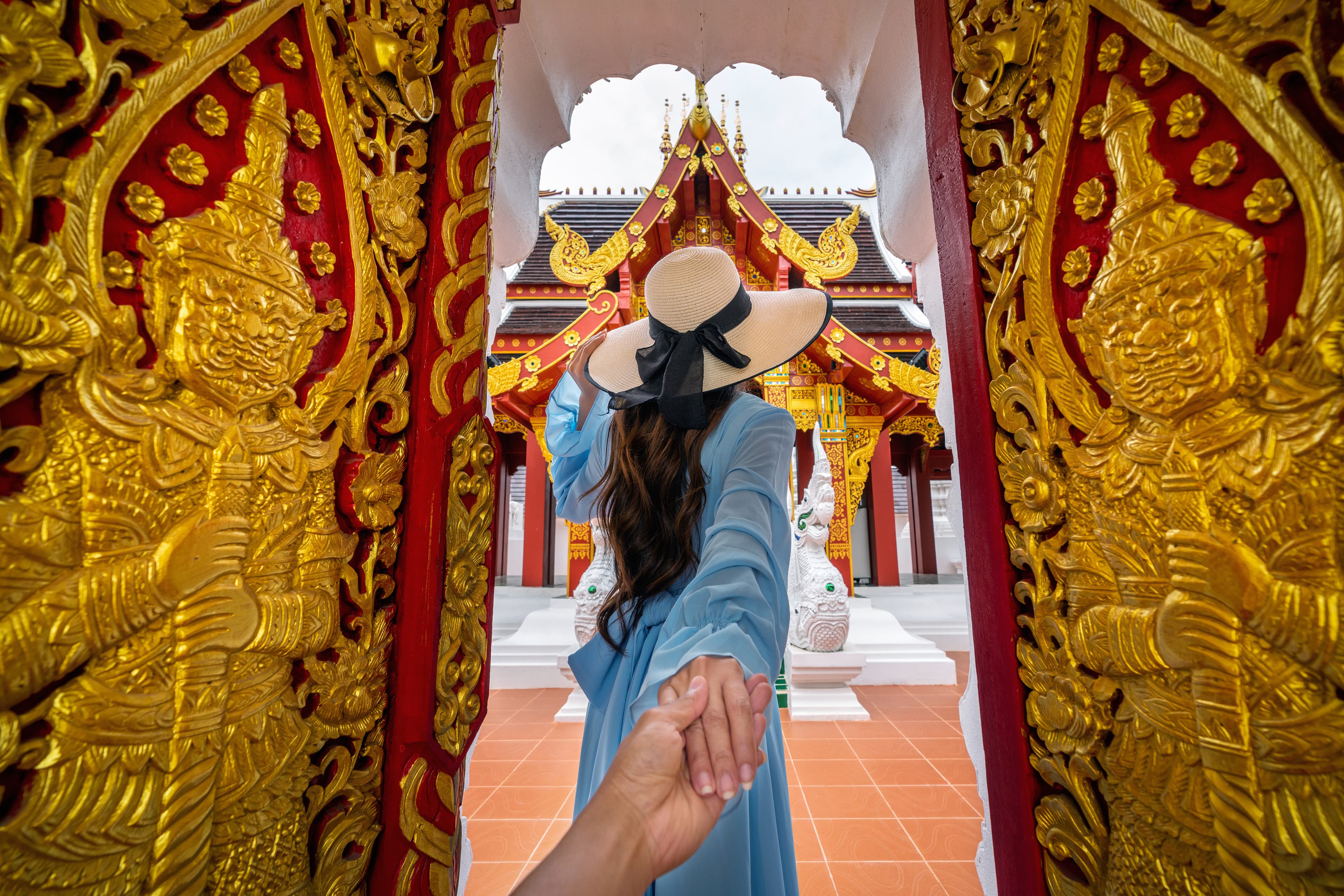 Woman in blue dress and straw hat leads someone through an ornate golden Thai temple.